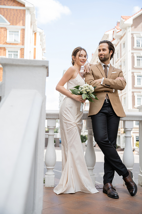 full length of happy young bride in wedding dress holding bouquet and leaning on groom in suit