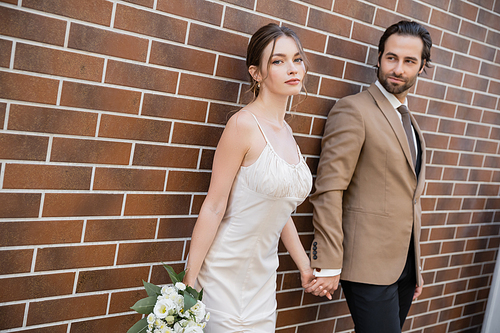 young bride in wedding dress holding bouquet and hand of groom near brick wall