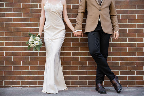 cropped view of young bride in white dress holding wedding bouquet and hand of groom near brick wall