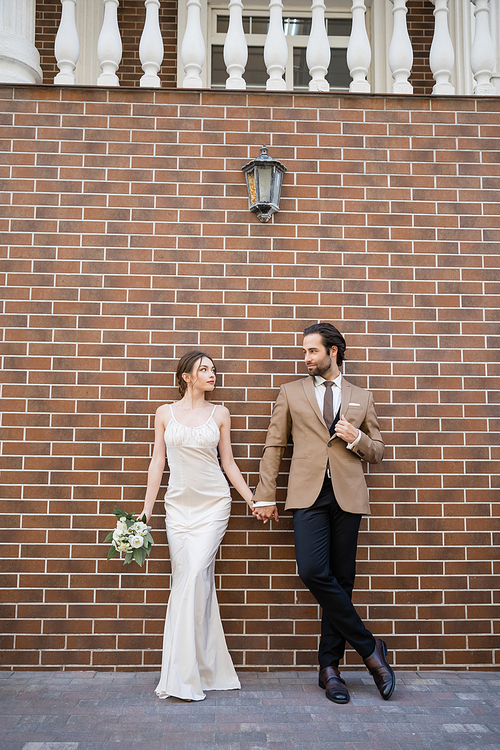 full length of bride in wedding dress holding flowers and hand of groom while standing against brick wall