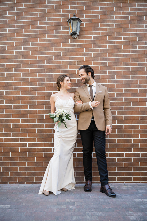 full length of happy bride in wedding dress holding flowers near groom in suit