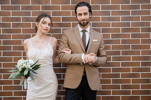 bride in wedding dress holding flowers near groom in suit while standing against brick wall