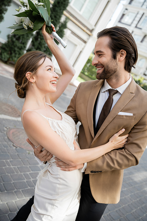 joyful bride in wedding dress holding bouquet and smiling with groom