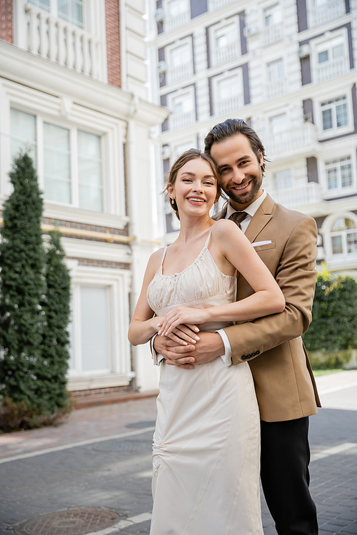 bearded man in suit with tie smiling and hugging happy bride in wedding dress