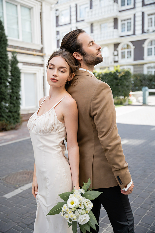 pretty bride in wedding dress holding bouquet and standing back to back with groom