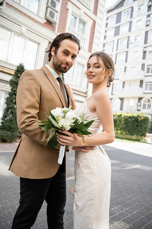 pretty bride in white wedding dress holding bouquet and smiling near groom