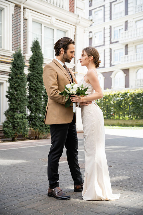 full length of pretty bride in white wedding dress holding bouquet and looking at groom