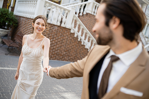 blurred groom in suit holding hand of happy bride in white dress