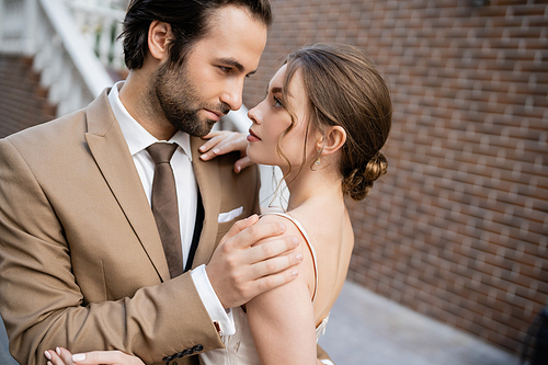 groom in beige suit looking at sensual bride in white dress while standing on street