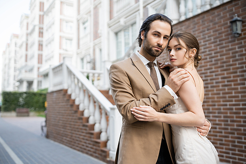 groom in beige suit hugging sensual bride in wedding dress while standing on street