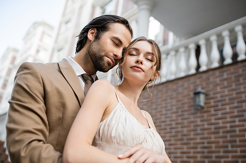 low angle view of groom in beige suit standing near gorgeous bride with closed eyes