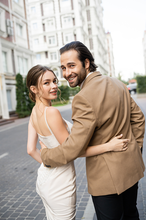 joyful groom in beige suit hugging happy bride in white wedding dress