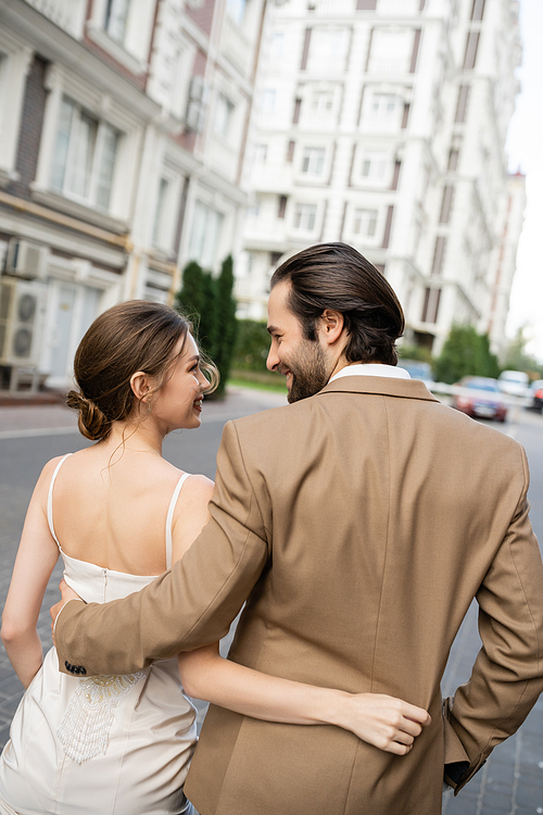 happy groom in beige suit hugging and looking at cheerful bride in white wedding dress