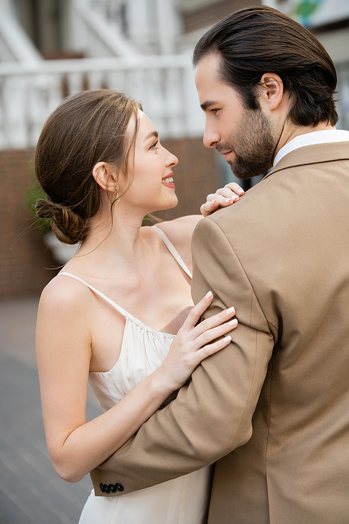 side view of groom in beige suit hugging smiling bride in white wedding dress