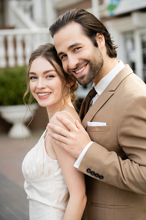 happy groom in beige blazer hugging gorgeous bride in white wedding dress