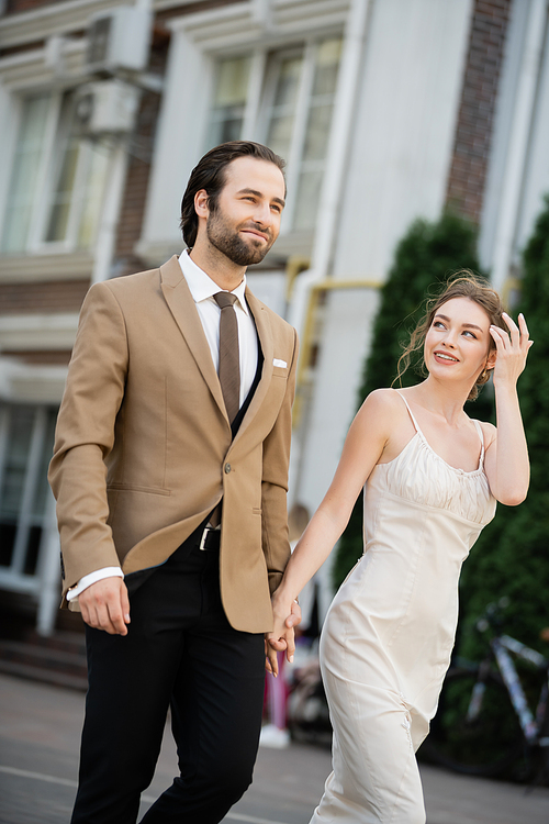 happy bride in wedding dress looking at groom while holding hands on street