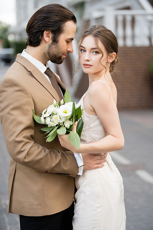 bearded groom hugging bride in wedding dress holding blooming flowers