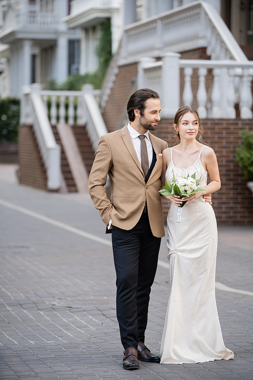 full length of groom posing with hand in pocket while looking at bride in wedding dress with blooming flowers