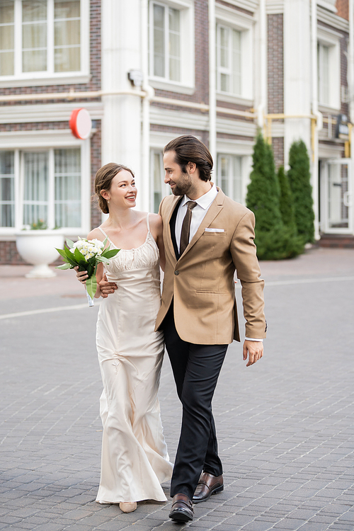 full length of happy bride in wedding dress holding blooming flowers and hugging with groom