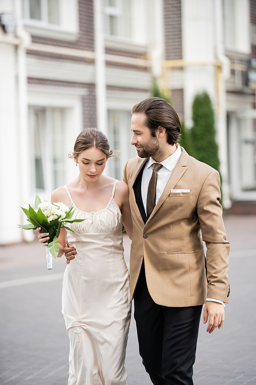 bride in wedding dress holding blooming flowers and hugging with bearded groom