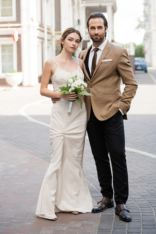 full length of bride in wedding dress holding blooming flowers near groom posing with hand in pocket