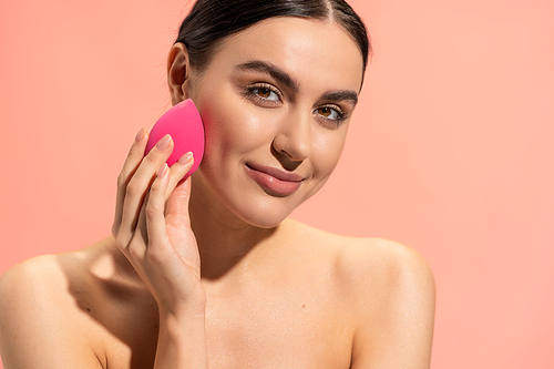 happy woman with bare shoulders applying face foundation with beauty sponge isolated on pink