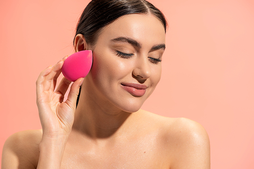 pleased woman applying face foundation with makeup sponge isolated on pink