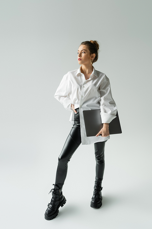 Full length of fashionable freelancer in shirt holding laptop and posing on grey