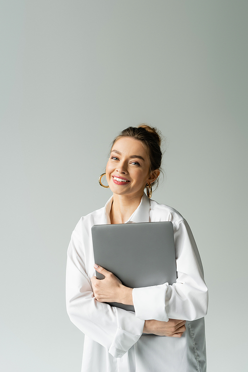 cheerful woman in white shirt holding laptop and looking at camera isolated on grey