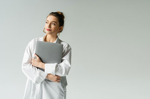 positive woman in white shirt holding laptop and looking away isolated on grey