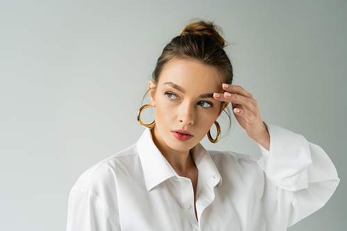 portrait of young woman in white shirt and hoop earrings touching eyebrow and looking away isolated on grey