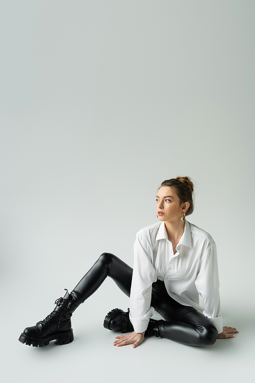 full length of trendy woman in white shirt and black tight pants sitting and looking away on grey background