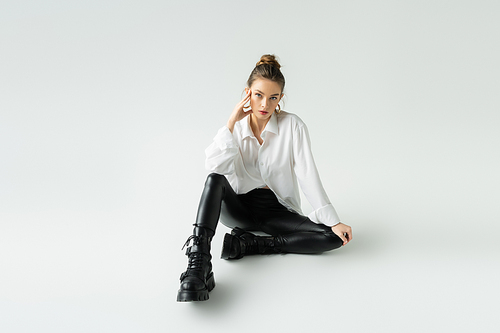 full length of young model in black latex pants and rough boots sitting on grey background