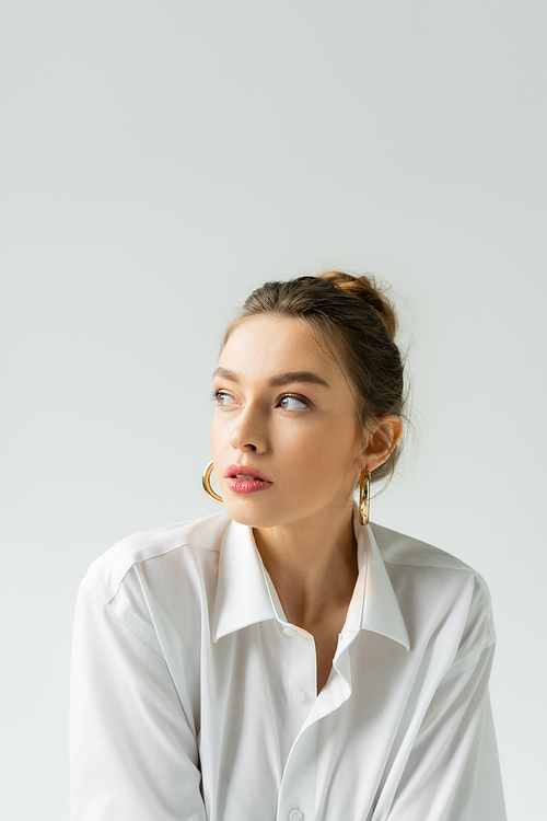 portrait of young woman in white shirt and hoop earrings looking away isolated on grey
