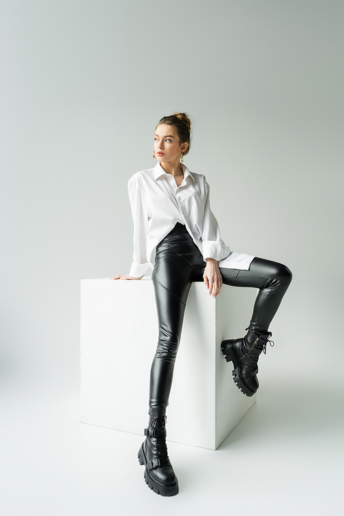 full length of model in white oversize shirt and black pants with rough boots sitting on white cube on grey background