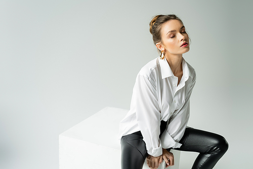 young sensual woman with closed eyes wearing white shirt and black latex pants and sitting on cube isolated on grey