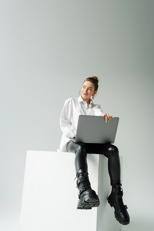 full length of happy woman in black latex pants and leather boots sitting on white cube with laptop on grey background