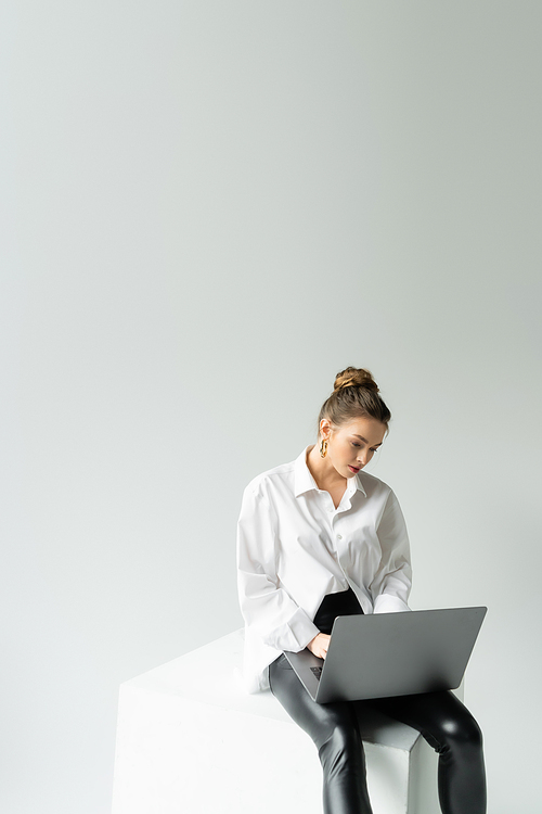 young woman in oversize shirt and black tight pants working on laptop while sitting on cube on grey background