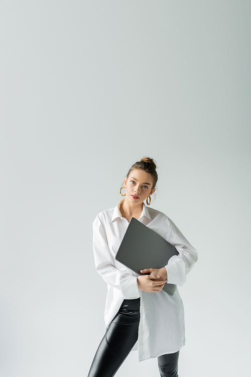 trendy woman in white shirt and hoop earrings posing with laptop and looking at camera isolated on grey
