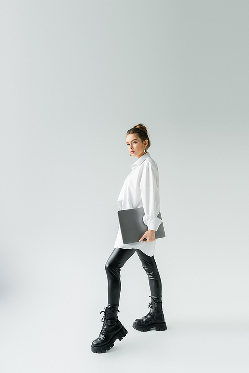 full length of stylish woman in black latex pants and rough boots posing with laptop on grey background