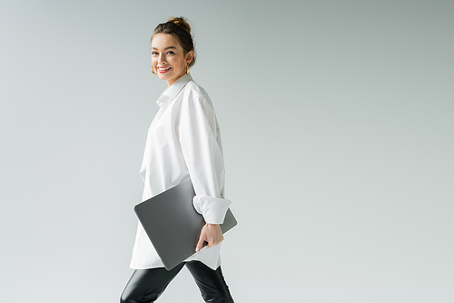 smiling woman in white shirt looking at camera while walking with laptop isolated on grey