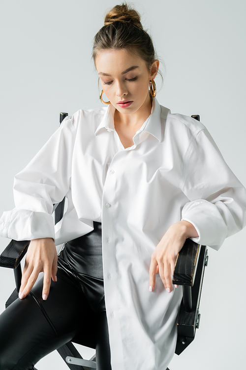 stylish woman sitting on chair in white oversize shirt isolated on grey