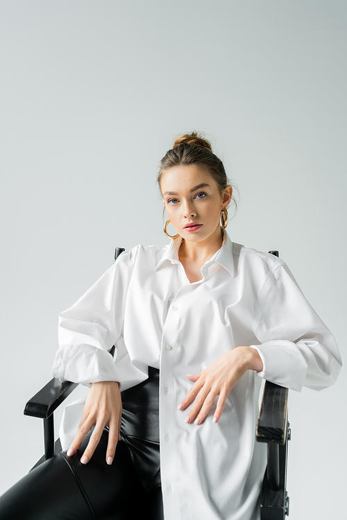 stylish woman in white shirt and golden earrings sitting on chair and looking at camera isolated on grey