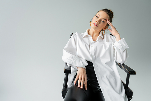 pretty woman in white oversize shirt and black tight pants posing on chair with hand near head isolated on grey