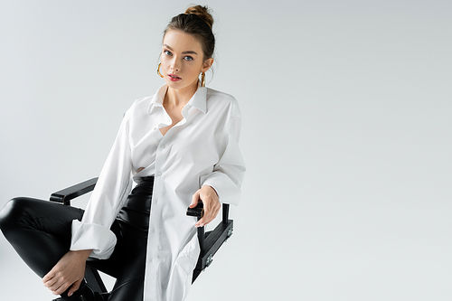 trendy woman in black tight pants and white oversize shirt sitting on chair and looking at camera isolated on grey