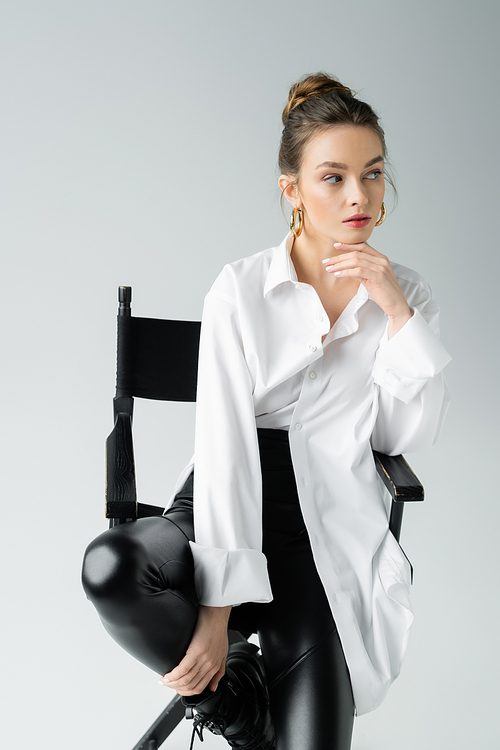 fashionable woman in white shirt and black latex pants sitting on chair and looking away isolated on grey