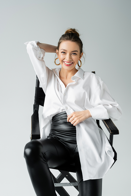 cheerful woman in white shirt and black latex pants smiling at camera while posing on chair isolated on grey