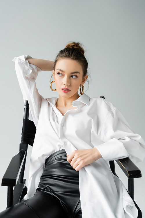 young model in white oversize shirt and black latex pants sitting on chair with hand behind head isolated on grey
