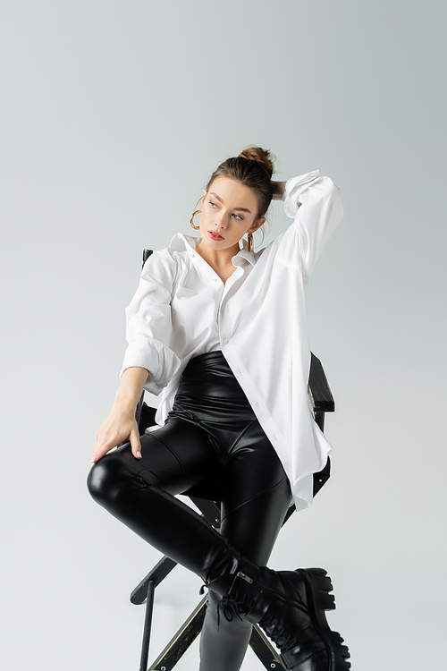 stylish woman in black tight pants and white shirt holding hand behind head while sitting on chair on grey background