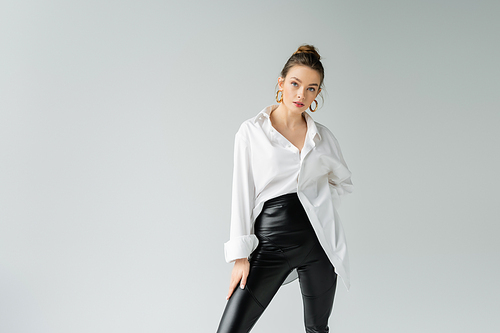 stylish woman in black tight pants and white oversize shirt posing and looking at camera isolated on grey
